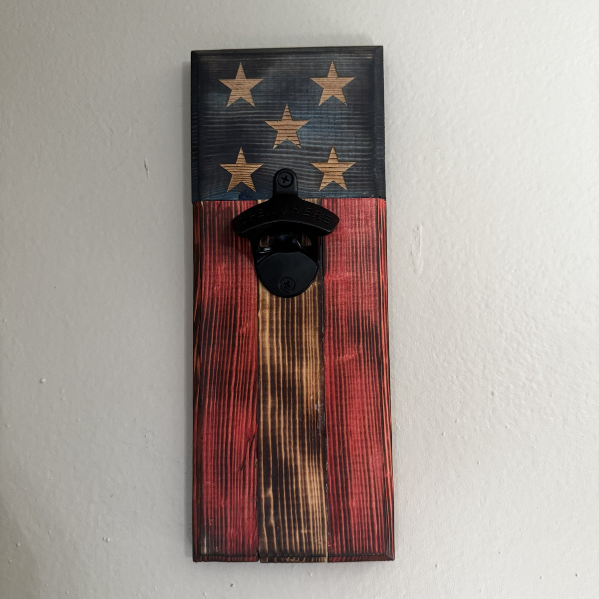 A rustic wooden wall-mounted bottle opener featuring a distressed American flag design with stars on a dark blue section and red, brown, and black wood stripes below, shown mounted on a white wall.
