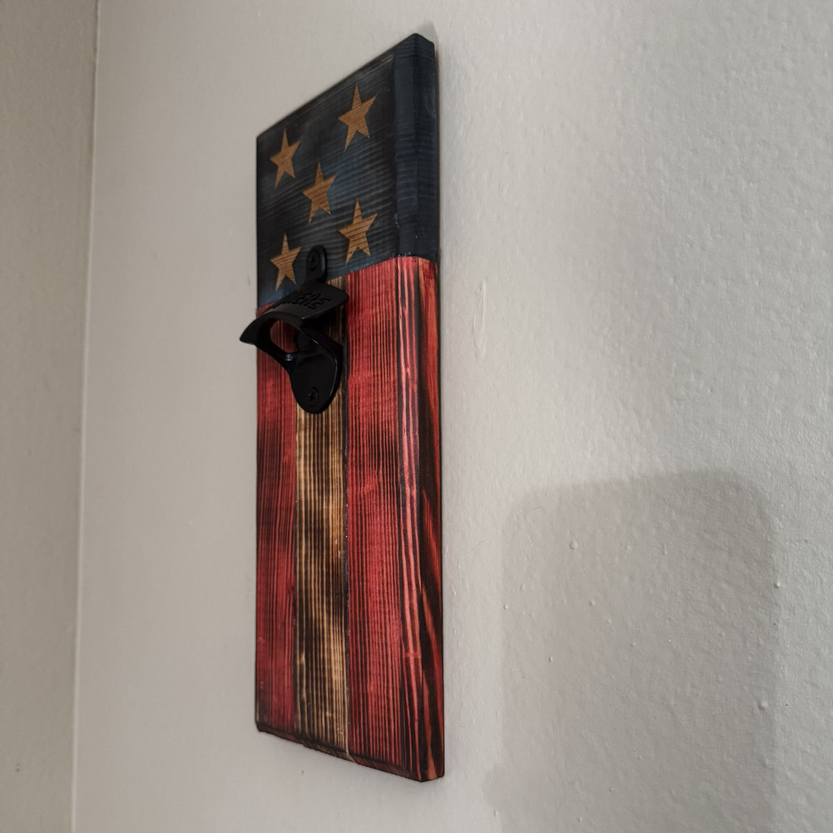 A rustic wooden wall-mounted bottle opener featuring a distressed American flag design with stars on a dark blue section and red, brown, and black wood grain stripes below, shown mounted on a white wall.