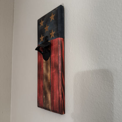 A rustic wooden wall-mounted bottle opener featuring a distressed American flag design with stars on a dark blue section and red, brown, and black wood grain stripes below, shown mounted on a white wall.
